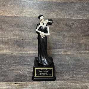 May include: A black and white skeleton couple figurine holding a bouquet of flowers. The figurine is on a black base with a gold plaque that reads "Best Couple Costume".