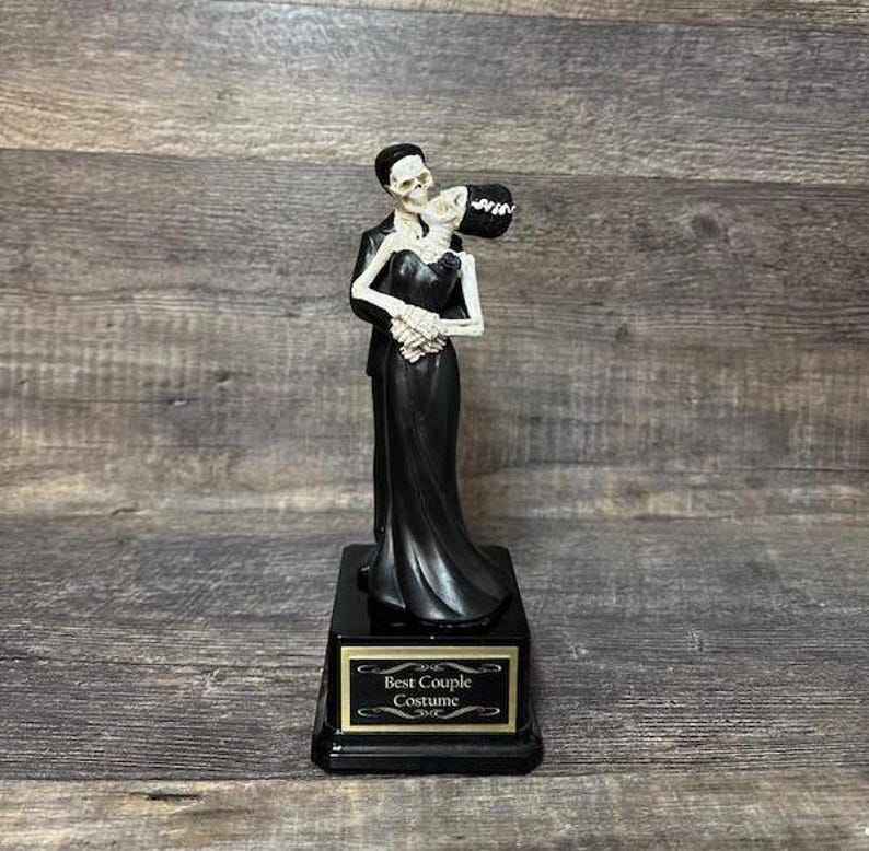 May include: A black and white skeleton couple figurine holding a bouquet of flowers. The figurine is on a black base with a gold plaque that reads "Best Couple Costume".
