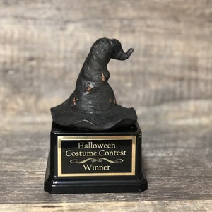 Op de afbeelding: Zwarte en gouden Halloween-kostuumwedstrijd winnaars trofee met een zwarte tovenaarshoed met gouden sterren. De trofee staat op een zwarte basis met een gouden plaquette waarop "Halloween-kostuumwedstrijd Winnaar" staat.