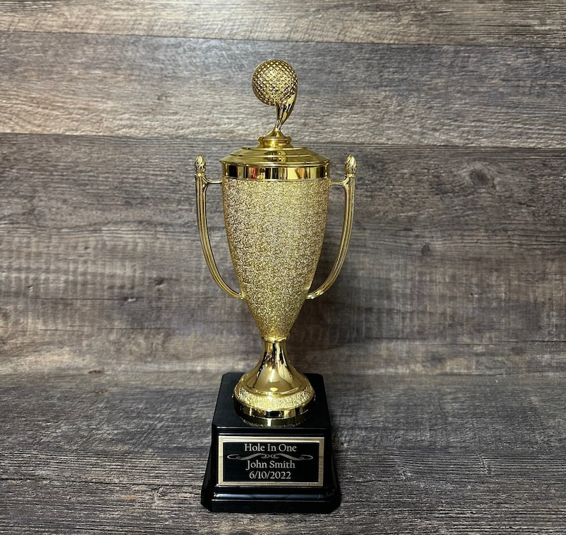 Może przedstawiać: Złoty puchar golfowy z piłką golfową na szczycie. Podstawa pucharu ma czarną tabliczkę z tekstem "Hole In One John Smith 6/10/2022".