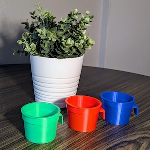 Puede incluir: Tres tazas de plástico de colores con asas. Las tazas son verde, roja y azul. Las tazas están sobre una mesa de madera.