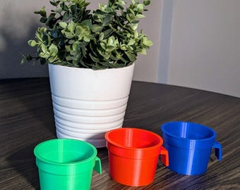 May include: Three colourful plastic cups with handles. The cups are green, red and blue. The cups are sitting on a wooden table.