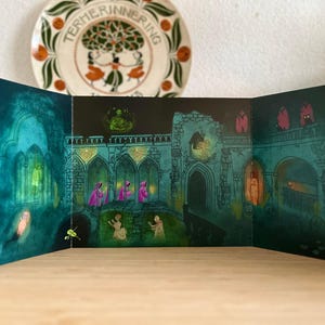May include: A tri-fold card featuring an illustrated fantasy scene with figures in pink robes, set in a teal-colored, gothic-style building. The artwork has a spooky, Halloween theme. A decorative plate is in the background.