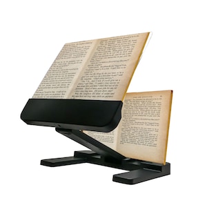 May include: A black adjustable book stand holding an open book with visible text. The stand has a base, an angled support, and a ledge to hold the book in place. The pages are yellowed, suggesting an older book.
