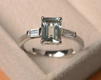 Op de afbeelding: Zilveren ring met een rechthoekige, lichtgroene edelsteen als middelpunt. Twee kleinere, witte baguette-geslepen stenen flankeren de hoofdsteen. De ring is geplaatst tegen een neutrale achtergrond.