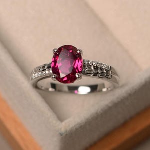 Könnte beinhalten: Ein silberner Ring mit einem großen, oval geschliffenen, tiefrosa Edelstein. Das Band ist mit kleineren, klaren Edelsteinen und aufwendigen Details verziert, was ein klassisches und elegantes Design ergibt.