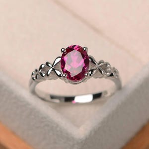 May include: A silver ring featuring an oval, magenta gemstone. The band has a floral design on each side of the gemstone. The ring is set against a soft, neutral background, highlighting the jewelry's details.
