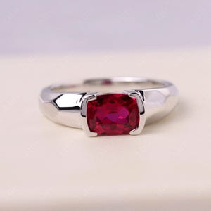 May include: A silver ring featuring a rectangular, deep red gemstone. The band has a modern, geometric design, and the gemstone is securely set in a bezel setting. The ring is displayed on a neutral background, highlighting its elegant simplicity.