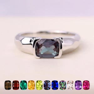 May include: A silver ring with a rectangular gemstone that changes colour. The gemstone displays teal, purple, and grey tones. A row of coloured gemstones is shown below the ring, displaying various hues.