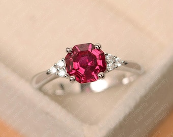 May include: A silver ring featuring a large, faceted, octagonal ruby gemstone as the centerpiece. Flanking the ruby are smaller, round, clear gemstones. The ring is set against a soft, neutral background.