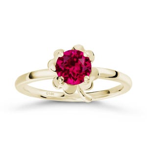 Op de afbeelding: Een gouden ring met een bloemvormige zetting met een helderrode edelsteen. De ring heeft een slanke band en een bloemmotief rond de centrale steen. De inscriptie "G14K" is zichtbaar op de band.