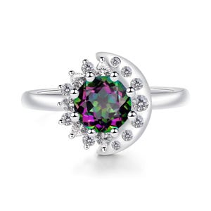 Op de afbeelding: Een zilverkleurige ring met een centrale, rond geslepen iriserende edelsteen met een veelkleurig oppervlak. De steen is omgeven door een halvemaanvormige zetting die langs de buitenrand en de basis is versierd met kleine, heldere, ronde stenen.