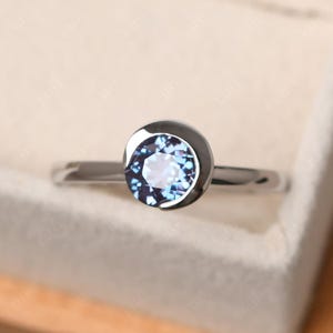 Puede incluir: Un anillo de plata con una piedra preciosa redonda, facetada, azul-púrpura engastada en un bisel. El anillo tiene una banda simple y pulida y un engaste oscuro que resalta el color de la piedra. Es una joya.