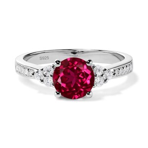 May include: A silver ring featuring a large, round, deep red gemstone. The ring is embellished with smaller, clear stones on either side and along the band. The inscription "S925" is visible on the band.