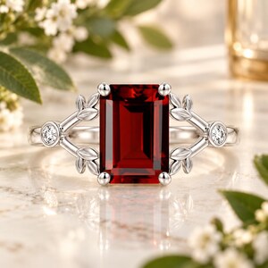 May include: A silver-toned ring featuring a large, rectangular, deep red gemstone at the center. The band is adorned with delicate metal leaf motifs on both sides, each accented by a small, round, clear gemstone set into the metal structure.