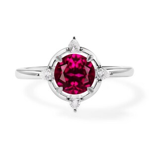 May include: A silver ring featuring a large, round, deep red gemstone at the center, surrounded by a halo of smaller white gemstones. The ring's band is thin and polished, with the gemstones set in a decorative pattern.
