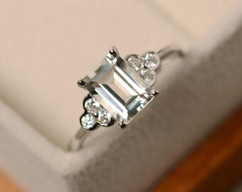Op de afbeelding: Een zilveren ring met een rechthoekige, lichtgroene edelsteen als middelpunt. De ring is versierd met kleinere, ronde, heldere edelstenen aan weerszijden van de hoofdsteen, waardoor een clusterontwerp ontstaat. De ring is gezet in een zilveren band.