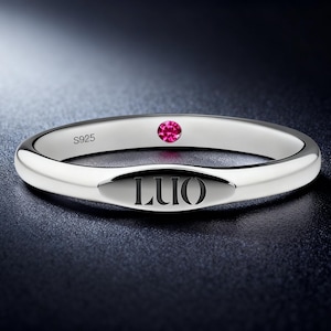 May include: A silver ring with a polished finish. The ring features a small, round, pink gemstone and the engraved word "LUO". The ring is marked with "S925".