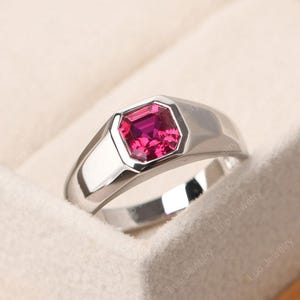 Puede incluir: Un anillo de plata con una llamativa gema magenta de corte cuadrado. El anillo tiene un diseño pulido y moderno con una banda ancha y un engaste de bisel. El anillo se muestra sobre una superficie blanca y suave.