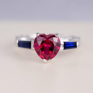Puede incluir: Un anillo de plata con una piedra preciosa roja en forma de corazón como pieza central. Dos gemas rectangulares de color azul oscuro flanquean el corazón. La banda del anillo es de plata pulida.
