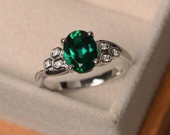 May include: A silver ring featuring a large, oval-cut emerald gemstone as the centerpiece. The ring is accented with smaller, round, clear gemstones on either side of the emerald. The ring is set in a silver band.