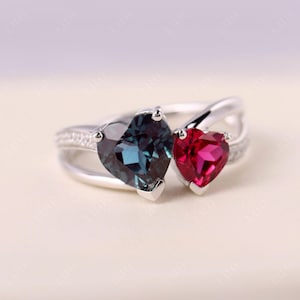 May include: A silver ring with two heart-shaped gemstones. One is a deep teal colour, and the other is a vibrant red. The ring band has a split design with small, clear stones set along one side, creating a romantic and elegant look.