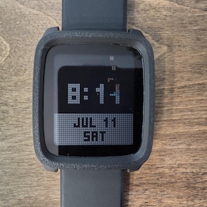 May include: A black digital smartwatch with a textured square frame and a silicone strap. The screen displays the time 8:14 in a pixelated font, with the date JUL 11 and the day SAT shown in a white rectangular box at the bottom of the display.