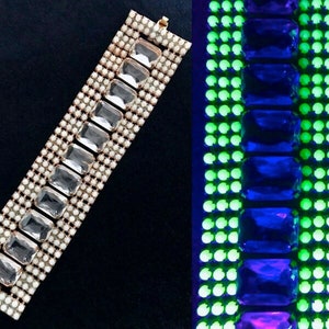 Puede incluir: Un brazalete vintage con brillantes rectangulares transparentes colocados en fila, rodeados de pequeños brillantes redondos blancos. El brazalete se muestra sobre un fondo negro. Un primer plano bajo luz negra revela un brillo verde neón.