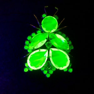 May include: A vibrant green insect-shaped brooch with a metallic gold frame. The brooch features large, faceted green gemstones arranged to resemble a beetle. The background is a dark, almost black color.