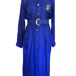 Könnte beinhalten: Ein kobaltblaues Kleid mit langen Ärmeln, passendem Gürtel und goldener Schnalle. Das Kleid hat eine Knopfleiste vorne, ein kleines gesticktes Emblem auf der Brust und ein rotes Kragenfutter. Das Kleid hat eine Midi-Länge.