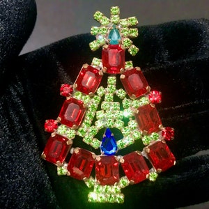 May include: A Christmas tree brooch with red, green, and blue gemstones. The brooch is shaped like a Christmas tree, with red rectangular gems forming the tree's body and green gems outlining the branches. A blue gem sits at the base.