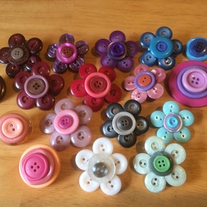 May include: Assortment of colourful flower-shaped buttons with various designs and colours, including pink, purple, blue, red, and grey.