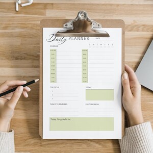 May include: A clipboard with a "Daily Planner" sheet, held by hands, with a pencil. The planner has a schedule, to-do list, and space for notes. A laptop and other office supplies are on the wooden desk.