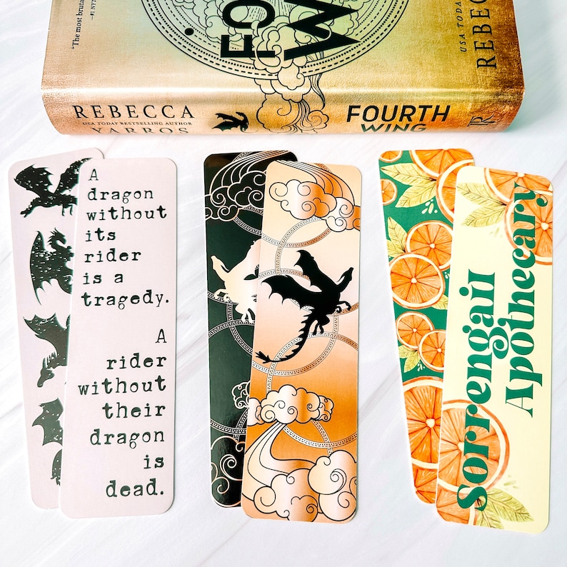 May include: A collection of decorative bookmarks with various designs. One bookmark features a dragon silhouette and text, "A dragon without its rider is a tragedy. A rider without their dragon is dead." Other bookmarks have dragon and orange slice designs.