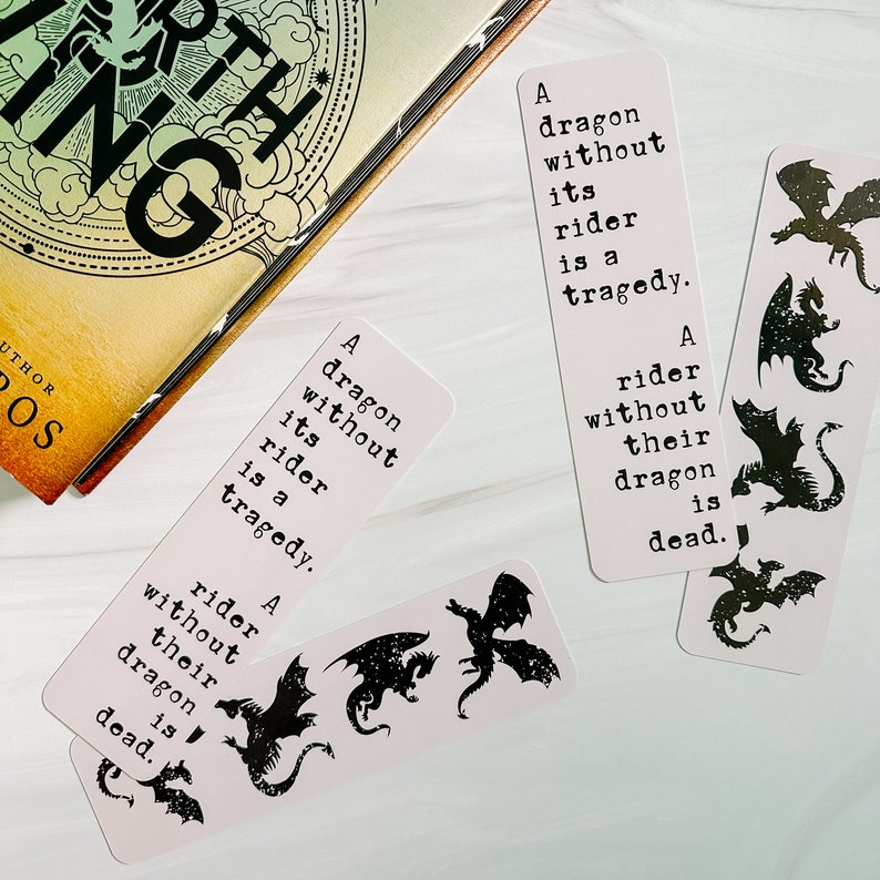 May include: Several white bookmarks with black dragon silhouettes and quotes from a fantasy novel. The text on the bookmarks reads, "A dragon without its rider is a tragedy. A rider without their dragon is dead."