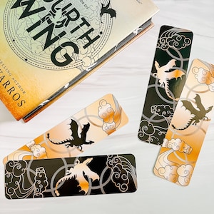 May include: Four dragon-themed bookmarks with a book. The bookmarks feature a dragon silhouette, clouds, and chain designs in black, gold, and orange. The book has the title "Fourth Wing" and the author's name.