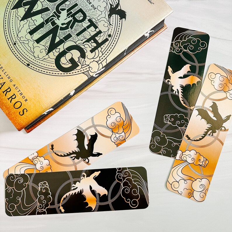 May include: Four dragon-themed bookmarks with a book. The bookmarks feature a dragon silhouette, clouds, and chain designs in black, gold, and orange. The book has the title "Fourth Wing" and the author's name.