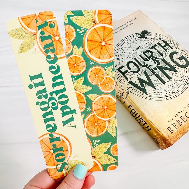 May include: Two decorative bookmarks with orange slice designs and the text "Sorrengail Apothecary" are next to a book titled "Fourth Wing". One bookmark has a cream background with orange slices and green text. The other bookmark has a teal background with orange slices and leaves.