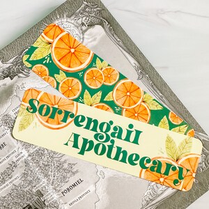 May include: Two decorative bookmarks with orange slice designs and the text "Sorrengait Apothecary." One bookmark has a green background with orange slices and leaves. The other has a cream background with the same design.
