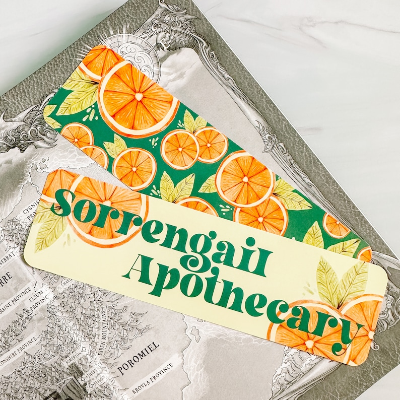 May include: Two decorative bookmarks with orange slice designs and the text "Sorrengait Apothecary." One bookmark has a green background with orange slices and leaves. The other has a cream background with the same design.