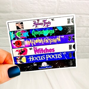 Puede incluir: Una pegatina con una pila de cintas VHS con títulos como "La Familia Addams", "Pesadillas", "Halloweentown", "Las Brujas" y "Hocus Pocus". La pegatina tiene un borde blanco y un aspecto vintage.