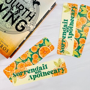 May include: Three bookmarks with an orange slice and leaf pattern, and the text "Sorrengail Apothecary" in green. A hardcover book with gold lettering is partially visible in the background.