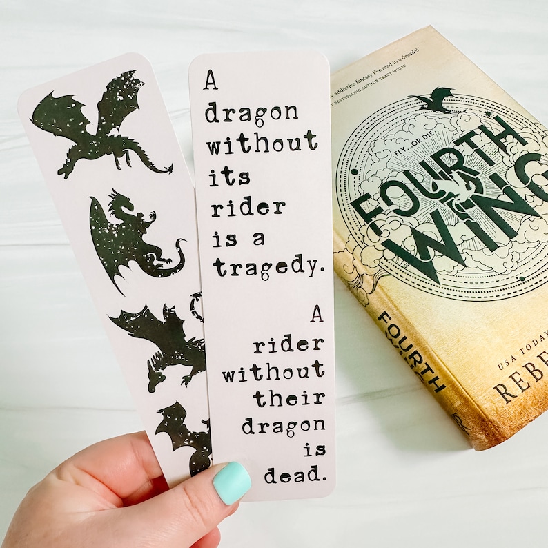 May include: Two light pink bookmarks with black dragon silhouettes and the quote: "A dragon without its rider is a tragedy. A rider without their dragon is dead." A book titled "Fourth Wing" is also visible.