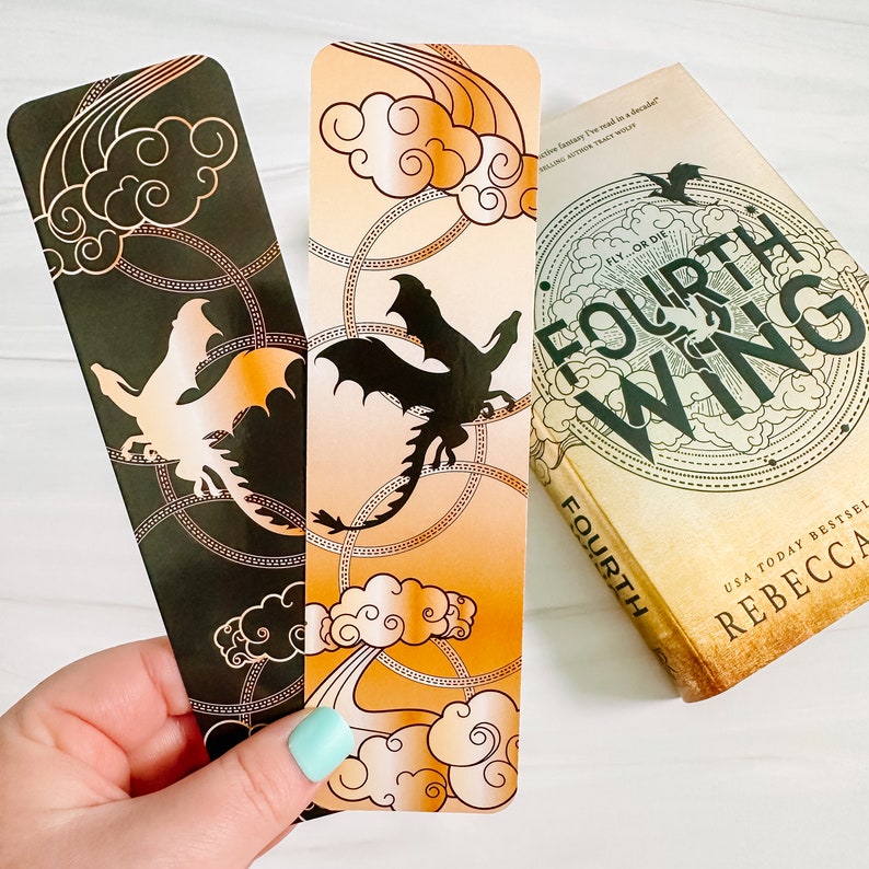 May include: Two dragon-themed bookmarks with gold accents and a book titled "Fourth Wing" by Rebecca. One bookmark is black with a gold dragon and cloud design. The other is orange with a black dragon and cloud design. The book has a circular design.