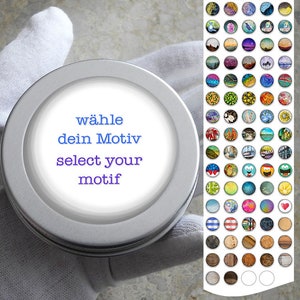 Puede incluir: Un recipiente redondo de plata con una etiqueta blanca que dice "wähle dein Motiv select your motif". El recipiente está sobre una superficie blanca. Hay una cuadrícula de imágenes redondas de colores en el fondo.