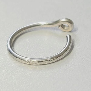 May include: A silver-colored nose ring with a hammered texture on the band. The ring has a curved design with a small loop at the top. The ring is shown on a light gray surface.