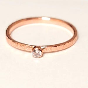 Könnte beinhalten: Ein roségoldener Ring mit strukturiertem Band und einem einzelnen, klaren Edelstein. Der Ring ist vor einem weißen Hintergrund platziert, der sein zartes Design hervorhebt. Der Edelstein ist klein und rund und verleiht ihm einen Hauch von Glanz.