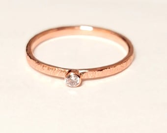 Puede incluir: Un anillo de oro rosa con una banda texturizada y una sola gema transparente. El anillo está sobre un fondo blanco, resaltando su delicado diseño. La gema es pequeña y redonda, agregando un toque de brillo.