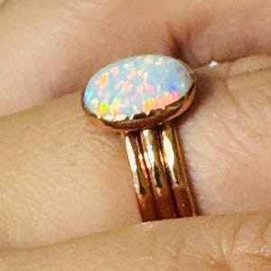 Op de afbeelding: Een roségouden ring met een ovale opaal edelsteen. De opaal toont een kleurenspel met iriserende flitsen van blauw, roze en geel. De ring heeft een drievoudig bandontwerp, wat bijdraagt aan de unieke esthetiek.