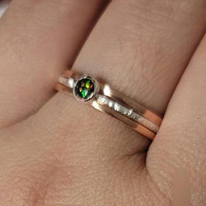 May include: A ring with a green and black opal gemstone set in a rose gold band. The ring is stacked with a silver band with a textured finish.